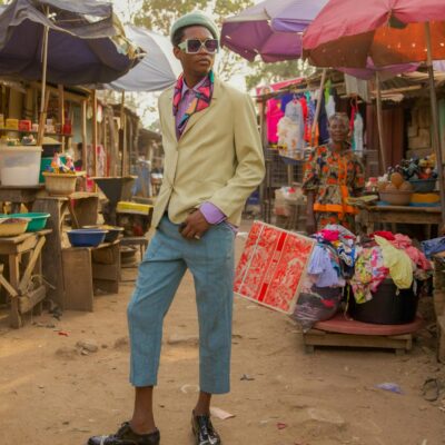 A fashionable man in colorful attire stands confidently in a bustling outdoor market.