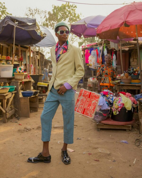 A fashionable man in colorful attire stands confidently in a bustling outdoor market.